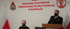 Na zdjęciu widoczny po prawej stronie przy mównicy w ubraniu wyjściowym przemawiający Komendant Powiatowy Państwowej Straży Pożarnej w Gorlicach bryg. Krzysztof Gładysz po jego lewej stronie w ubraniu dowódczo sztabowym Naczelnik Wydziału ds. Operacyjno-szkoleniowych w Gorlicach. Strażacy na tle białej ściany z godłem i z napisem koloru czerwonego Komenda Powiatowa Państwowej Straży Pożarnej w Gorlicach po bokach flagi biało czerwone. 
