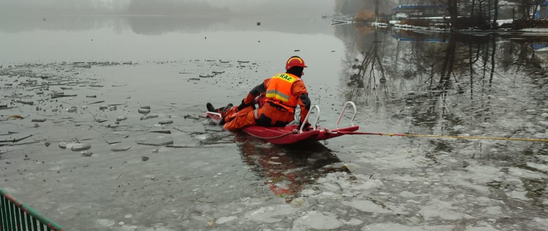 Zdjęcie przedstawia akwen jeziora, częściowo pokryty skruszonym lodem. Na jego powierzchni jeden człowiek ubrany w pomarańczowo-czarny kombinezon i czerwony kask przemieszcza się na czerwonych saniach lodowych asekurując leżącego innego człowieka. W tle przybrzeżne drzewa.