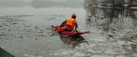 Zdjęcie przedstawia akwen jeziora, częściowo pokryty skruszonym lodem. Na jego powierzchni jeden człowiek ubrany w pomarańczowo-czarny kombinezon i czerwony kask przemieszcza się na czerwonych saniach lodowych asekurując leżącego innego człowieka. W tle przybrzeżne drzewa.