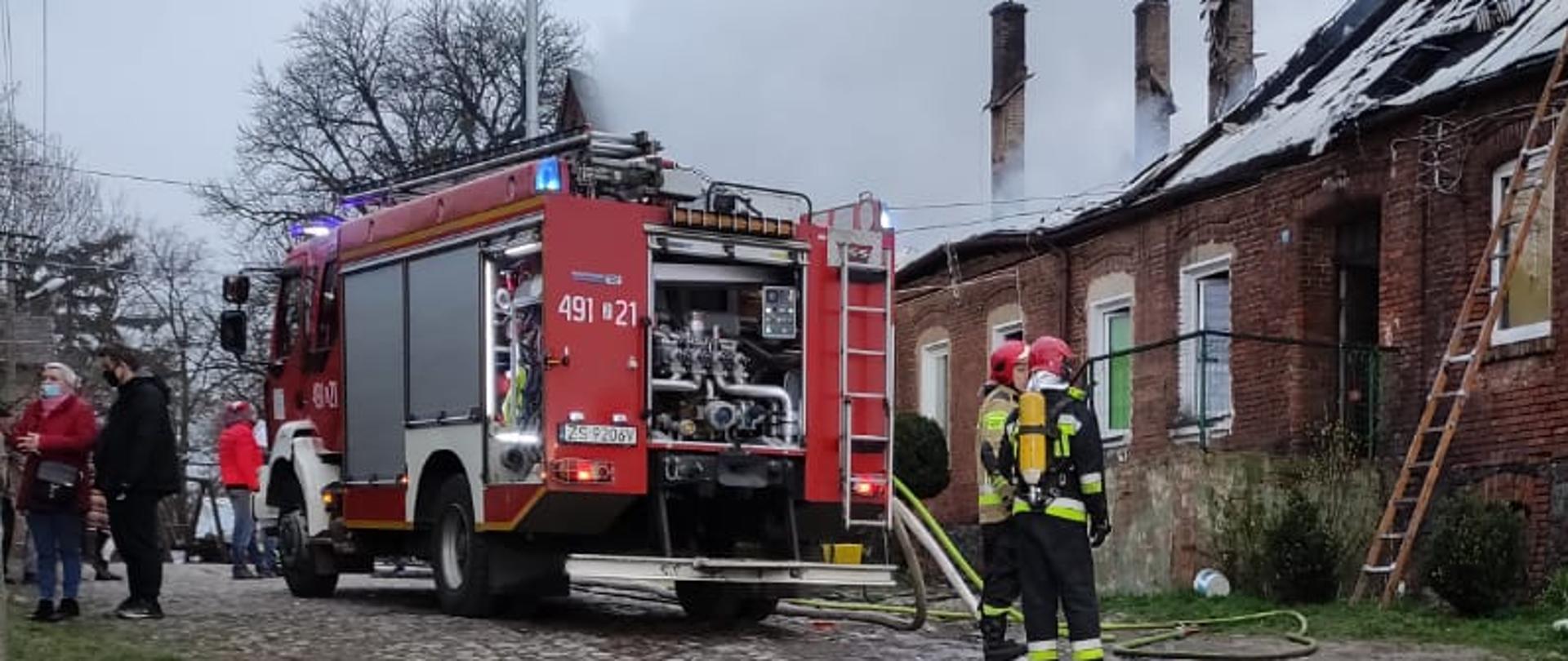 Na zdjęciu strażacy ubrani w umundurowanie bojowe i hełmy stoją obok budynku z którego wydobywa się dym. W tle pojazd pożarniczy i gapie.