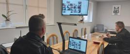 Zdjęcie przedstawia dwóch strażaków w czarnych mundurach dowódczo – sztabowych siedzących w pomieszczeniu przy drewnianym biurku i stole. Mężczyźni biorą udział w wideokonferencji śledząc ja na zamontowanym na ścianie telewizorze i stojącym na biurku komputerze przenośnym.