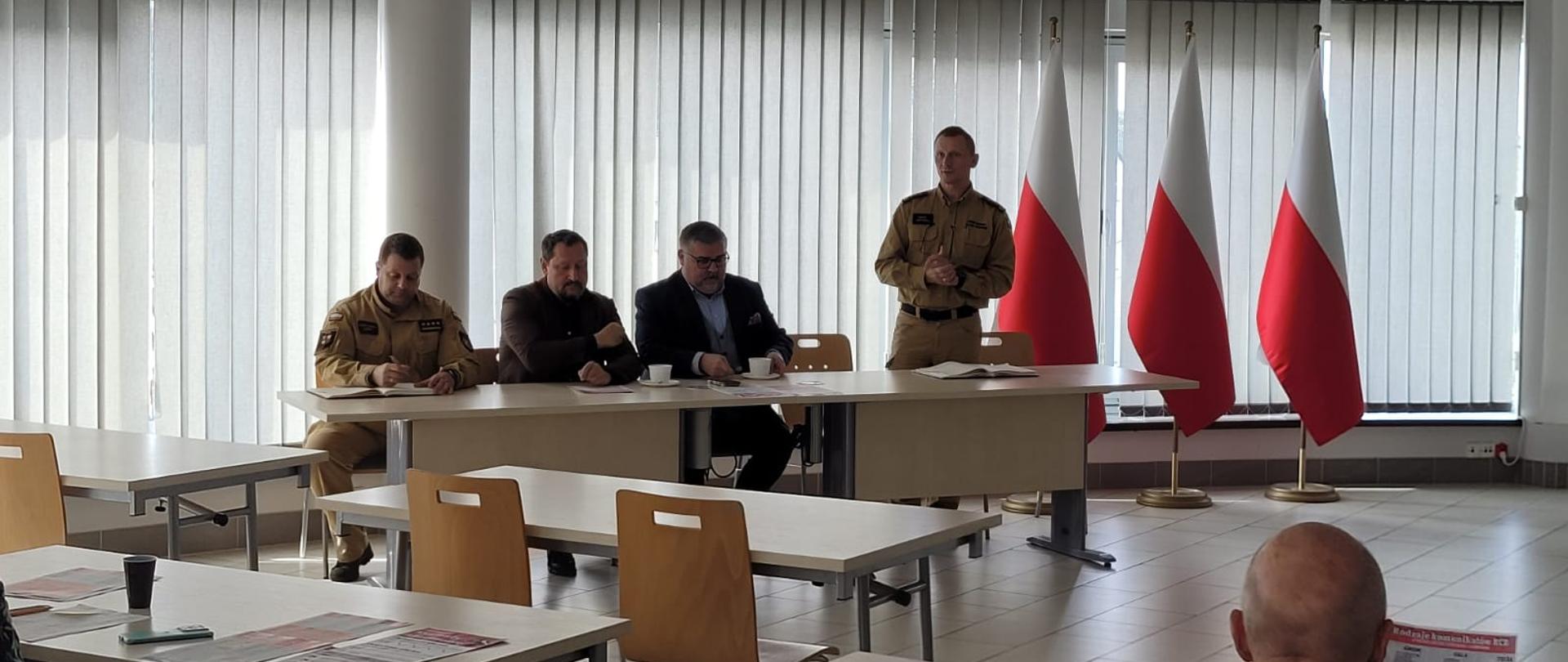 Jasna sala szkoleniowo-konferencyjna. Przy stole ustawionym przed oknami z żaluzjami i trzema stojącymi flagami Polski przemawia stojący funkcjonariusz Państwowej Straży Pożarnej w piaskowym mundurze służbowym. Obok niego przy stole siedzi inny umundurowany strażak oraz dwóch mężczyzn w garniturach. Na pierwszym planie widoczne są puste i zajęte stoliki uczestników spotkania.