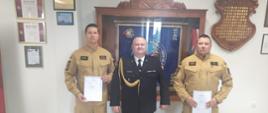 Strażacy stoją na tle gabloty ze sztandarem. W środku u mundurze galowym stoi komendant a po jego lewej i prawej stronie strażacy z mianowani na wyższe stanowiska służbowe.