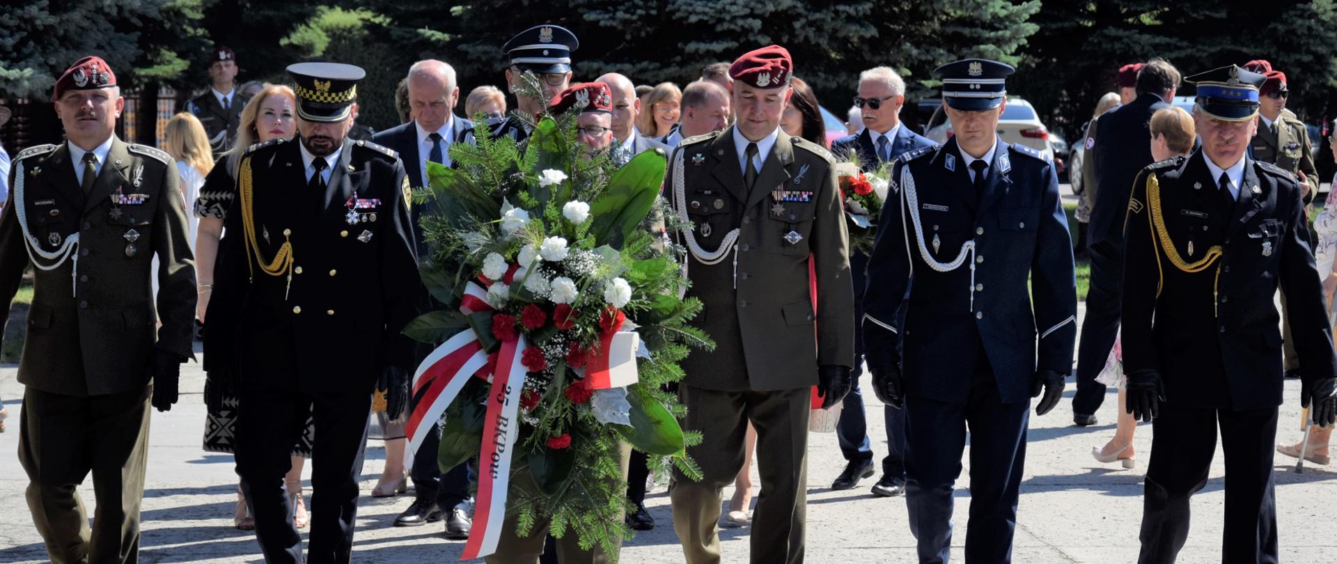 Na zdjęciu widać funkcjonariuszy ze straży pożarnej, policji, wojska, straży miejskiej. Jeden z żołnierzy niesie wiązankę, która przedstawiciele służ mundurowych złożą przed grobem nieznanego żołnierza.