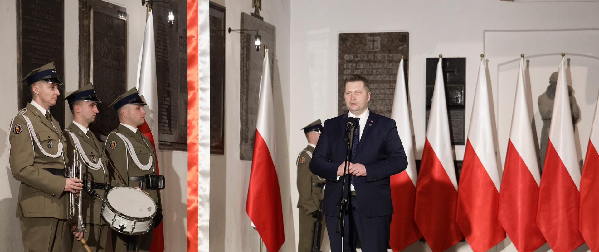 Minister Przemysław Czarnek przemawia przy mikrofonie. W tle biało-czerwone flagi i liczne tablice zawieszone na ścianach. Po prawej stronie żołnierze ubrani w galowe mundury. Jeden z nich trzyma w ręku trąbkę, drugi biały bębenek. 