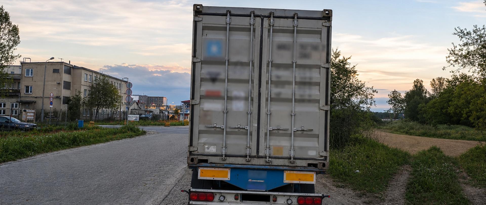 Fotografia przedstawia tył nieprawidłowo zaparkowanej ciężarówki z kontenerem, stojącej przy bocznej drodze