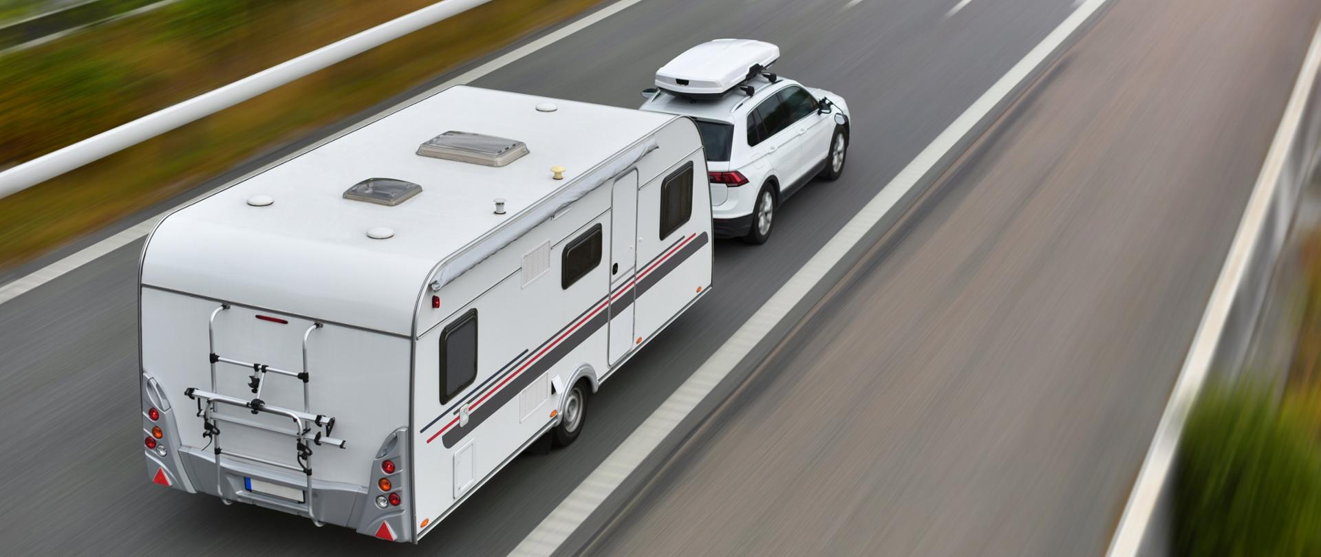 Biały samochód z przyczepą kempingową jedzie drogą ekspresową, widziany z góry z rozmytym tłem.
