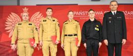 Pięć osób w mundurach strażackich stoi w szeregu na tle czerwonej ścianki z symbolem orła i napisem Państwowa Straż Pożarna.
