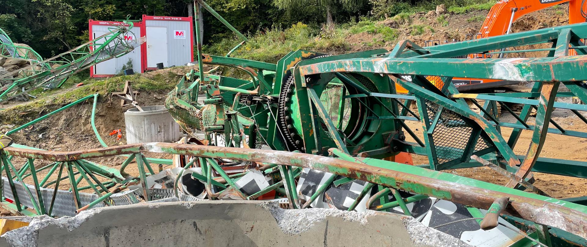 Na zdjęciu widać przewrócony żuraw. Żuraw jest koloru zielonego. Zdjęcie jest wykonane w dzień. Żuraw leży na skraju skarpy obok budowanego budynku. 