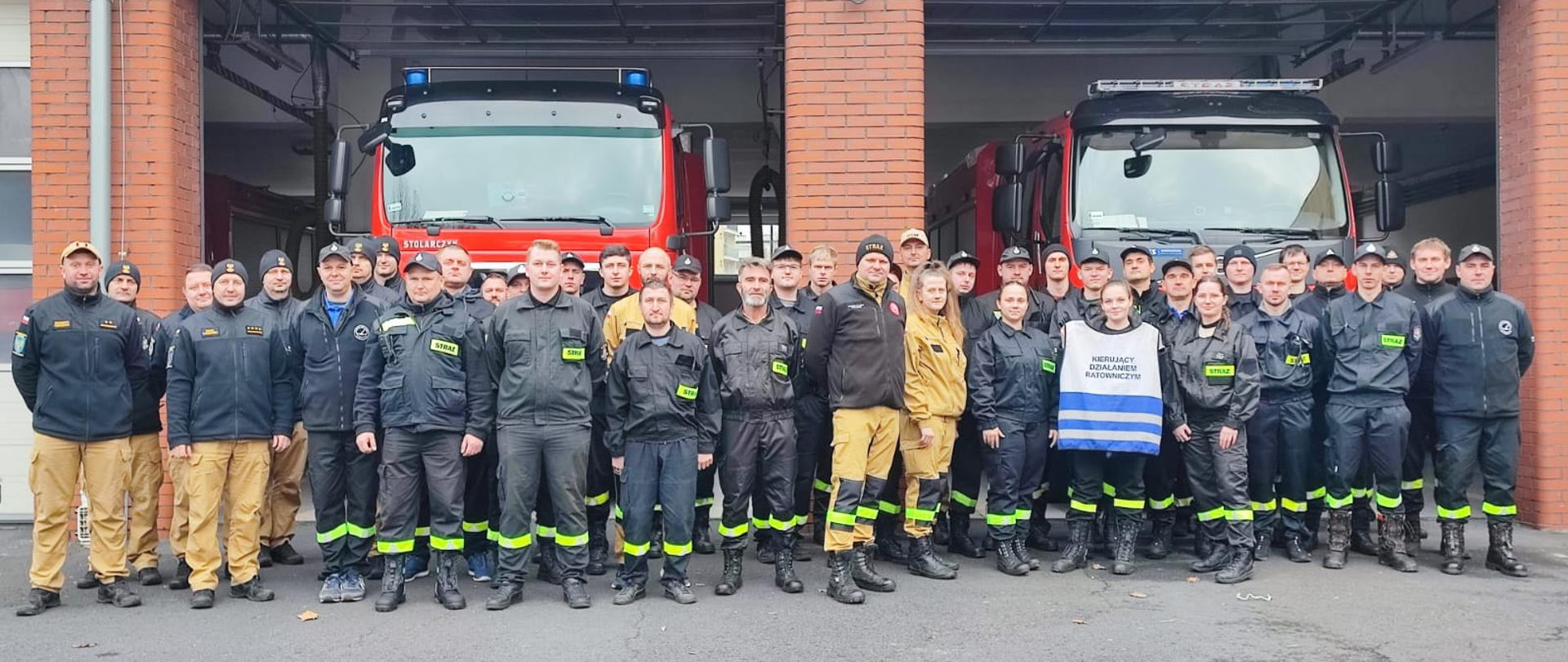 Na zdjęciu widać druhów OSP, funkcjonariusza KW PSP w Opolu oraz funkcjonariuszy KP PSP w Oleśnie. Wszyscy w ubraniach koszarowych na tle samochodów pożarniczych i garażu Komendy Powiatowej PSP w Oleśnie. Jedna z osób trzyma w ręku kamizelkę Kierującego Działaniem Ratowniczym.