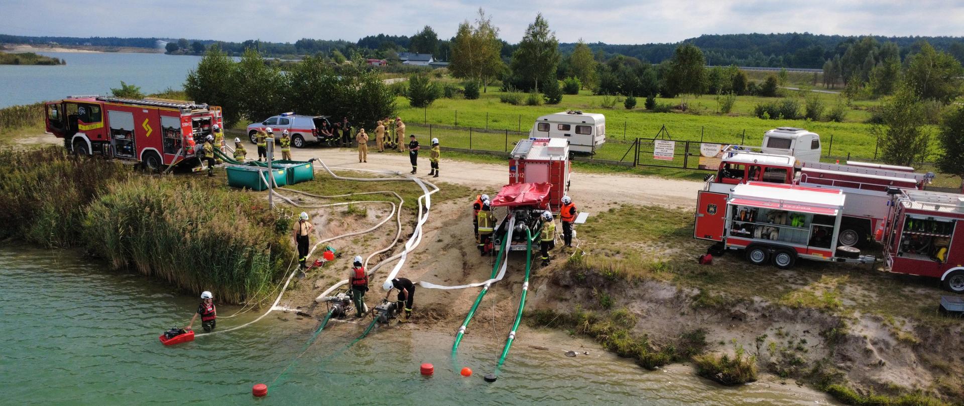 Samochody , pompy pożarnicze węze i zbiornik wodny ustawone dad brzegiem zbiornika podają wodę 