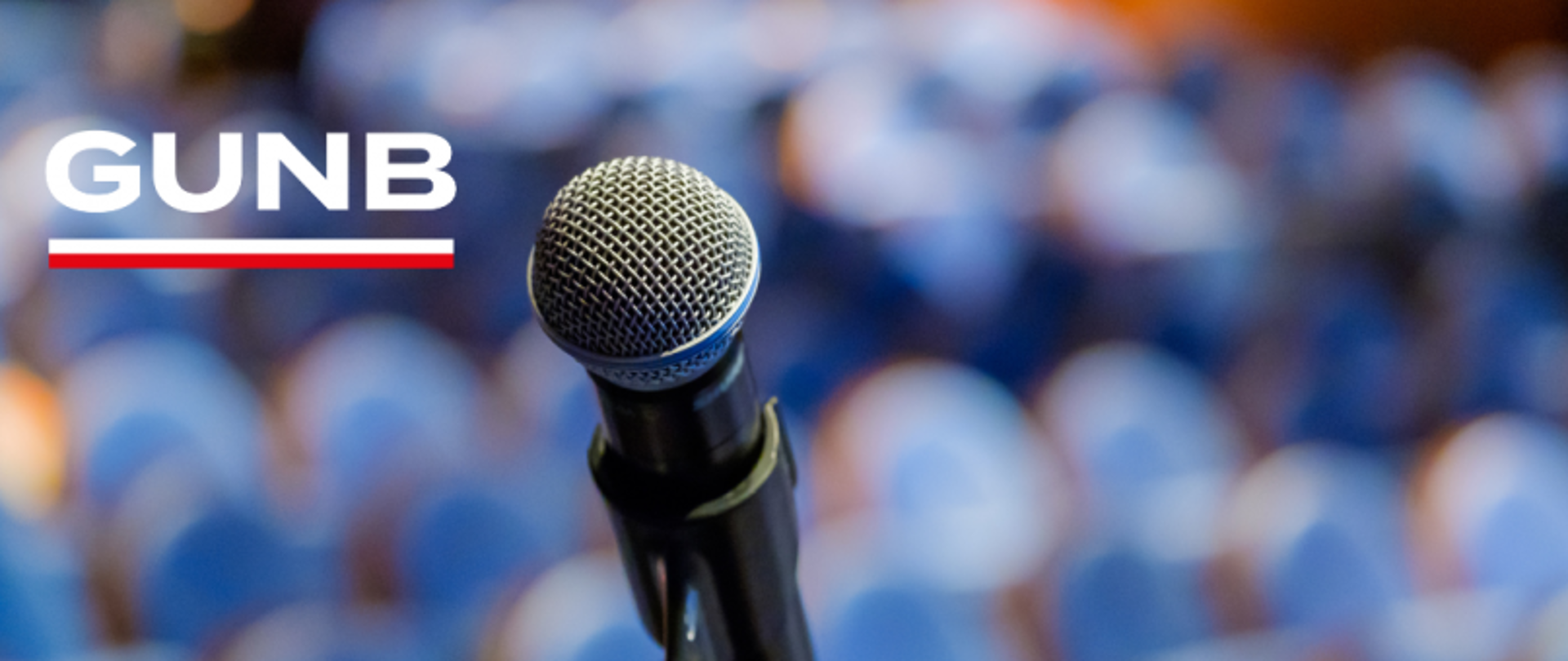 Mikrofon na tle rozmytej sali konferencyjnej z logo GUNB w lewym górnym rogu.