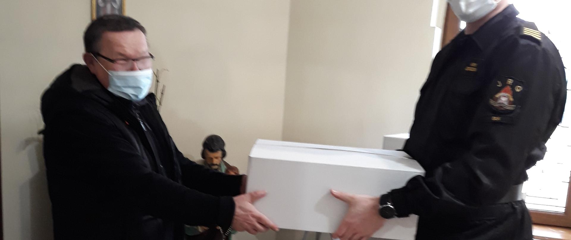Na zdjęciu znajduje się strażak, który przekazuje maseczki proboszczowi w pomieszczeniach kościoła