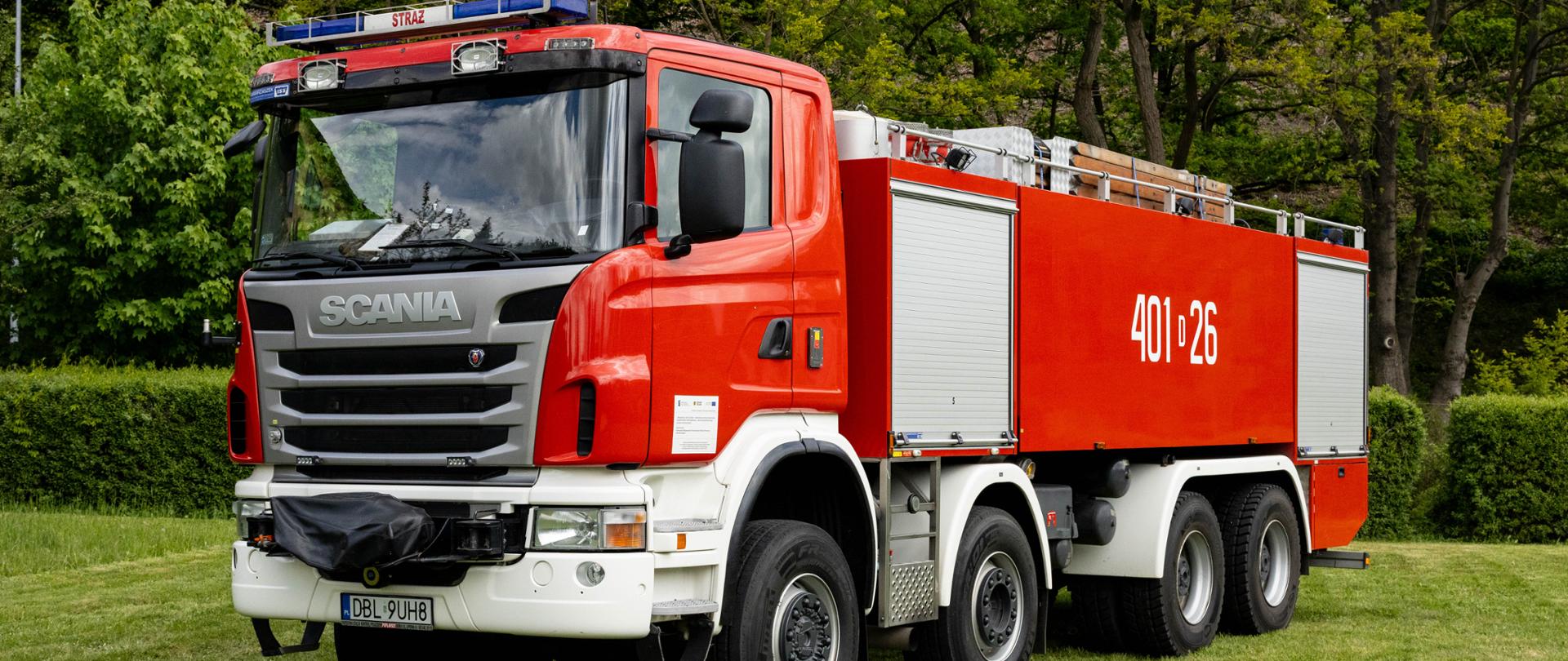 Ciężki, czteroosiowy samochód gaśniczy Scania G480 w charakterystycznym czerwonym kolorze z białymi błotnikami. Na dużym, gładkim boku zabudowy umieszczono biały numer operacyjny „401[D]26”. Na dachu widoczna jest m.in. drabina pożarnicza.