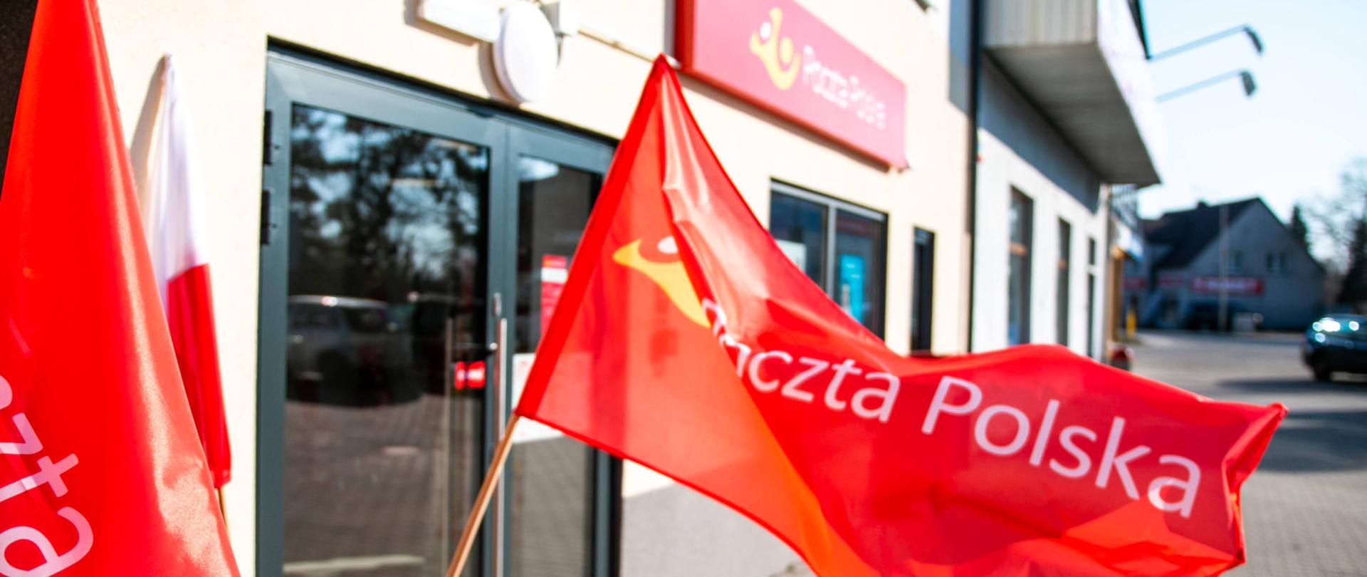 Budynek Poczty Polskiej z dużymi szklanymi drzwiami. Na pierwszym planie powiewa czerwona flaga z napisem "Poczta Polska". W tle domy. 