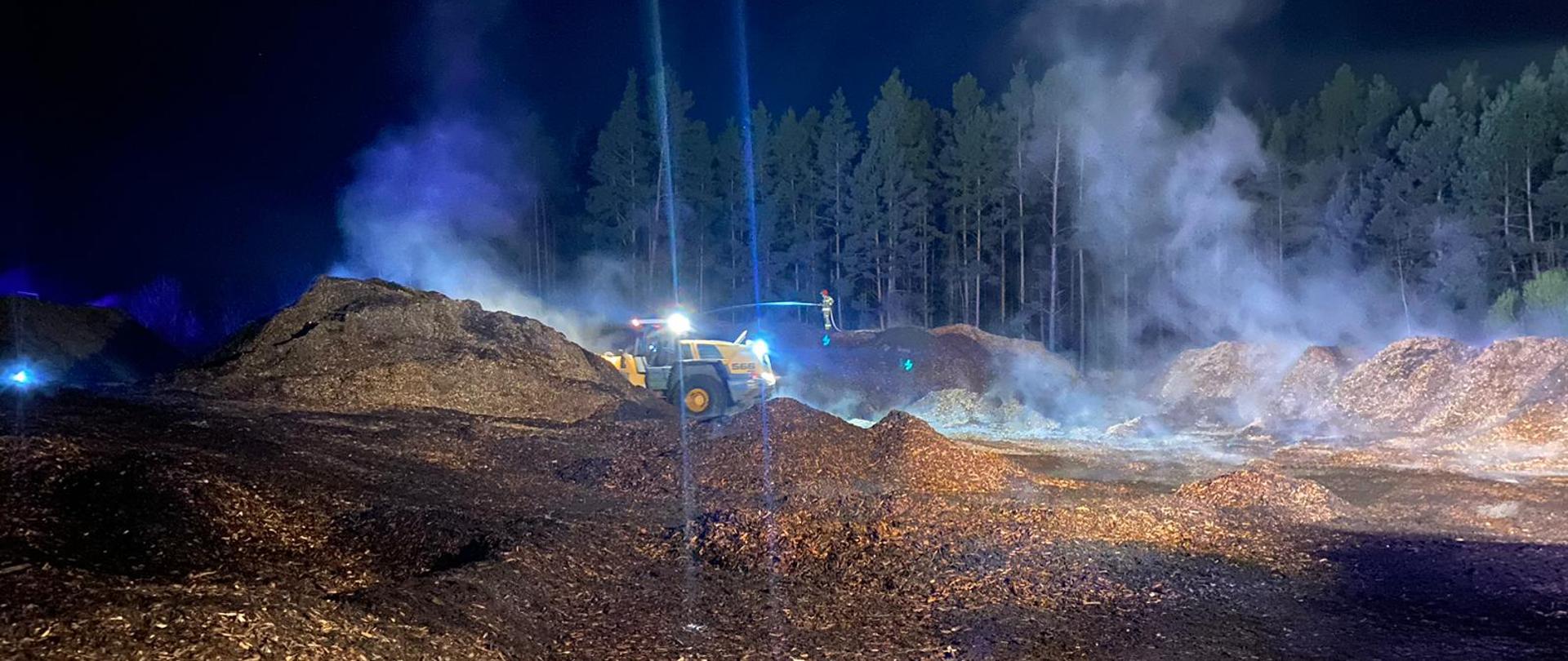 Na hałdach zrębki stoi żółta ładowarka, która przerzuca zrębkę. Ze zrębki wydobywa się dym. 