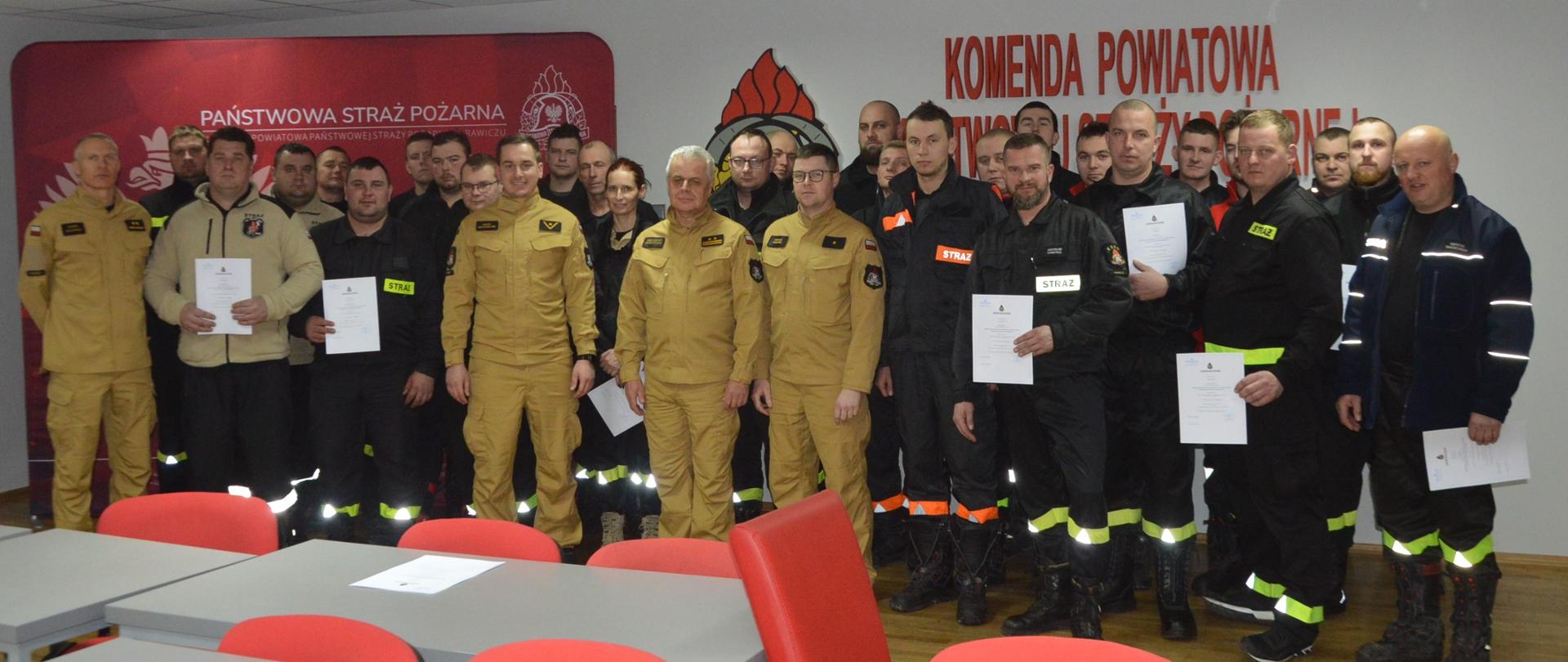 Egzamin KDR. Świetlica w Komendzie Powiatowej PSP w Rawiczu. Zdjęcie grupowe uczestników szkolenia wraz z komisją egzaminacyjną i zastępcą komendanta powiatowego PSP. Strażacy stoją na tle ściany z napisem KP PSP i logiem PSP oraz ścianki promującej KP PSP w Rawiczu.