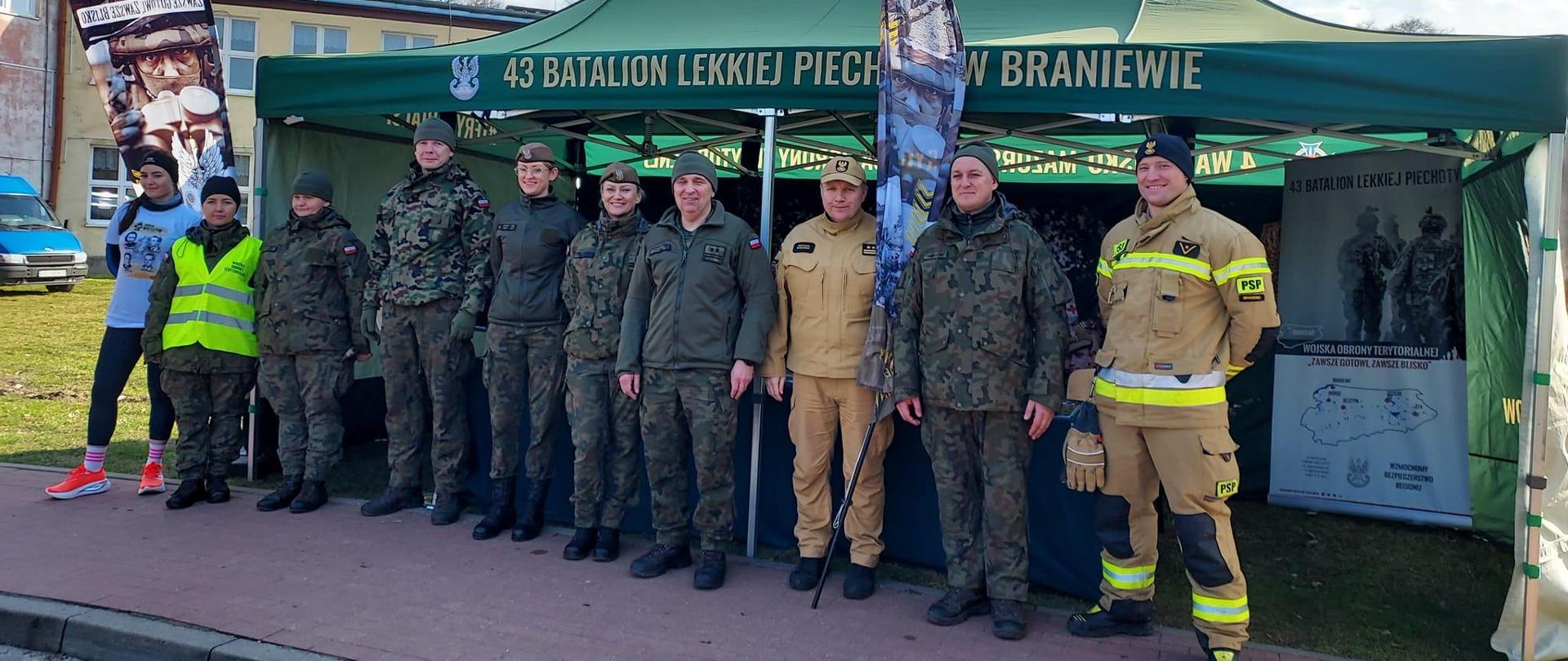 Pod zielonym namiotem, stoją przedstawiciele służb mundurowych w mundurach. Słonecznie.