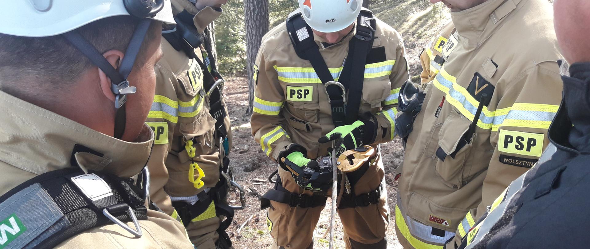 Grupa strażaków zapoznaje się z budową oraz zasadą działania sprzętu do ratownictwa wysokościowego. Jeden z nich, ubrany w szelki bezpieczeństwa przygotowuje się do zejścia w dół na linie.