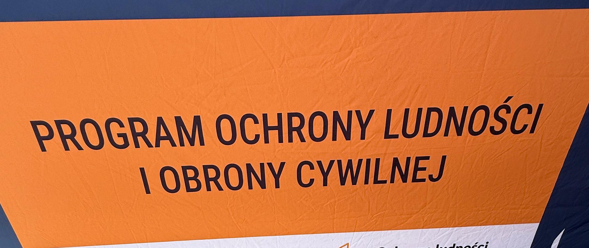 Zdjęcie przedstawia stoisko informacyjne Ministerstwa Spraw Wewnętrznych i Administracji (MSWiA) poświęcone Programowi Ochrony Ludności i Obrony Cywilnej.
Na tle stoiska znajduje się duży baner w barwach pomarańczowej, białej i ciemnoniebieskiej. Pośrodku widnieje wielki napis wielkimi literami: „PROGRAM OCHRONY LUDNOŚCI I OBRONY CYWILNEJ”. W lewym dolnym rogu znajduje się orzeł w koronie (godło Polski) oraz mniejszy napis „Ministerstwo Spraw Wewnętrznych i Administracji”. W prawym dolnym rogu umieszczono logo i napis: „Ochrona ludności i obrona cywilna”.
Na stole ustawionym przed banerem leży czarny plecak z widocznymi czerwonymi napisami: „APTECZKA” (umieszczonym nad mniejszymi białymi napisami: „SPODNIE/POGOTOWIE”) i „NARZĘDZIA”. Obok plecaka, po lewej stronie, ułożone są w kolumnach białe koperty, z których górna ma napis „Poradnik bezpieczeństwa”.
Na środku stołu znajdują się rzędy mniejszych białych karteczek, na których wielokrotnie powtórzony jest napis w trzech linijkach: „PLECAK EWAKUACYJNY”. Po prawej stronie tych kartek znajdują się z kolei mniejsze białe ulotki z czarnymi napisami i wieloma malutkimi rysunkami, prawdopodobnie przedstawiającymi wyposażenie plecaka ewakuacyjnego.
Zdjęcie wykonano w świetle dziennym, a na stole widać cienie.