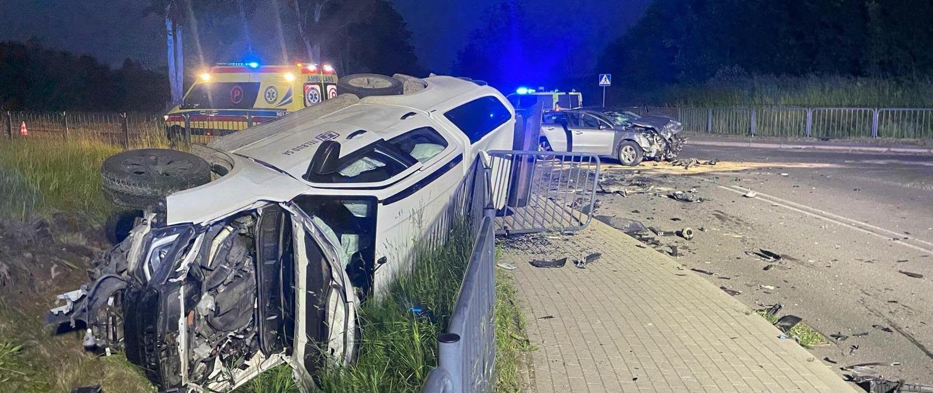 Zderzenie dwóch pojazdów osobowych. jedn z samochodów z lewej strony lezy na boku poza drogą, połamał barierki ochronne. Drugi w oddali w poprzek drogi. Widać światła ostrzegawcze służb. Pora nocna.