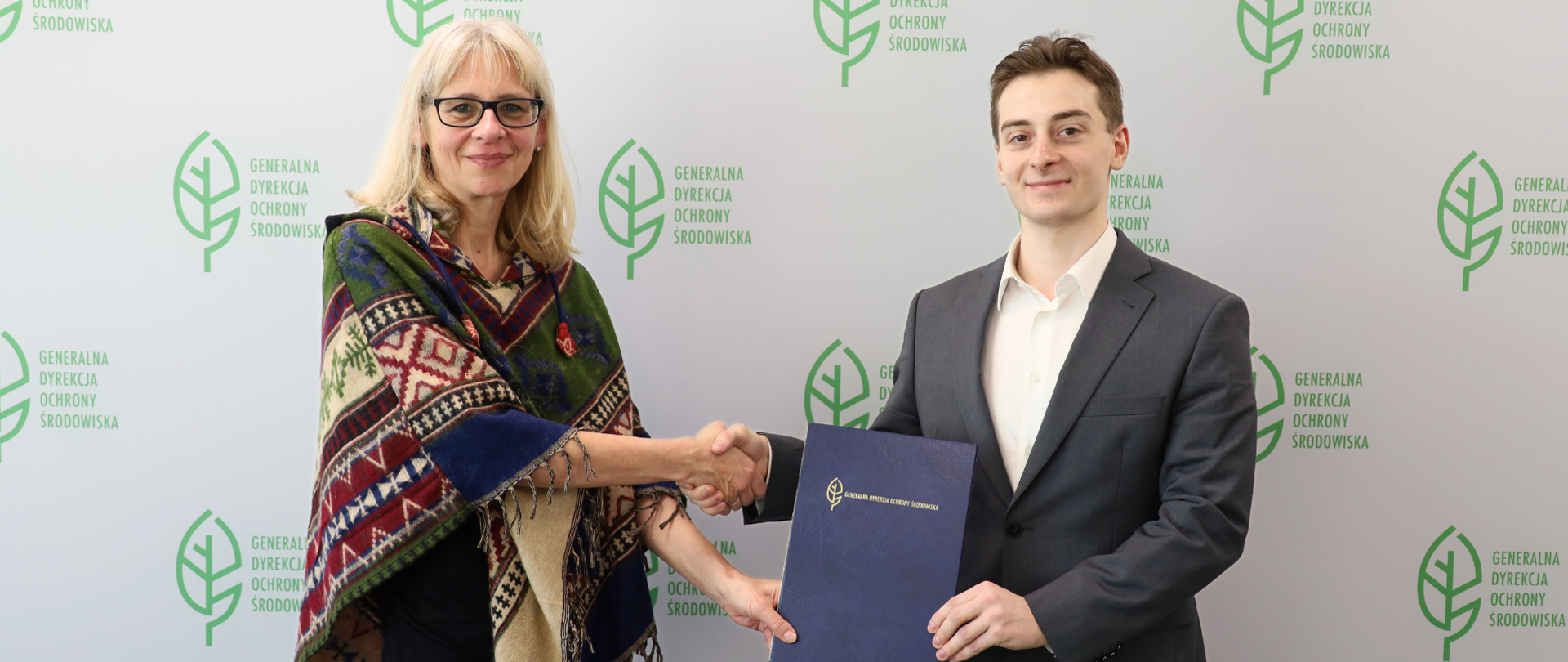 Na tle białej ścianki z logo Generalnej Dyrekcji Ochrony Środowiska (zielony liść) stoją kobieta i mężczyzna, którzy ściskają sobie dłonie. Drugimi dłońmi trzymają teczkę.