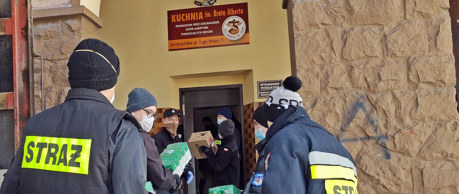 Ośmiu strażaków w ubraniach koszarowych, czapkach oraz maseczkach i rękawiczkach ochronnych wyładowuje z samochodu ciężarowego paczki z ryżem i papierem toaletowym. Na pierwszym planie paczki z puszkami z pasztetem drobiowym oraz uchwyt wózka rozładowczego. W tle widoczne wejście do budynku, na którym znajduje się tablica z napisem „KUCHNIA św. Brata Alberta PROWADZONA PRZEZ ZGROMADZENIE SIÓSTR ALBERTYNEK POSŁUGUJĄCYCH UBOGIM, Andrychów ul. 1-go Maja 11”
