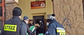 Ośmiu strażaków w ubraniach koszarowych, czapkach oraz maseczkach i rękawiczkach ochronnych wyładowuje z samochodu ciężarowego paczki z ryżem i papierem toaletowym. W tle widoczne wejście do budynku, na którym znajduje się tablica z napisem „KUCHNIA św. Brata Alberta PROWADZONA PRZEZ ZGROMADZENIE SIÓSTR ALBERTYNEK POSŁUGUJĄCYCH UBOGIM, Andrychów ul. 1-go Maja 11”