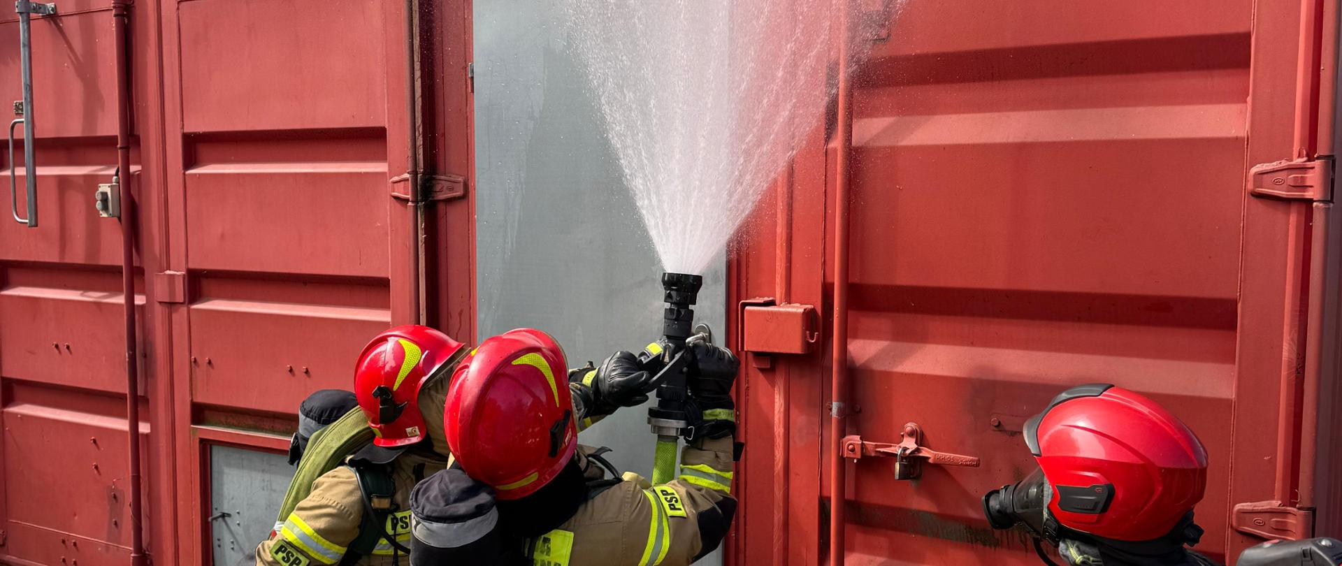 Strażacy Państwowej Straży Pożarnej na terenie poligonu ratowniczego Komendy Wojewódzkiej PSP w Opolu w umundurowaniu bojowym ćwiczą działania związane z pożarami wewnętrznymi. Strażacy ćwiczą wejście z prądem wody gaśniczej do zadymionej komory rozgorzeniowej. Komora rozgorzeniowa zbudowana na bazie kontenerów.