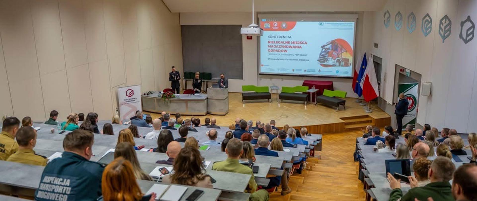 Aula akademicka. Widok z góry na rzędy miejsc siedzących, na sali kilkadziesiąt osób. W dole stół, mównica i trzy osoby. Z boku widać kanapy, za nimi duży ekran. 