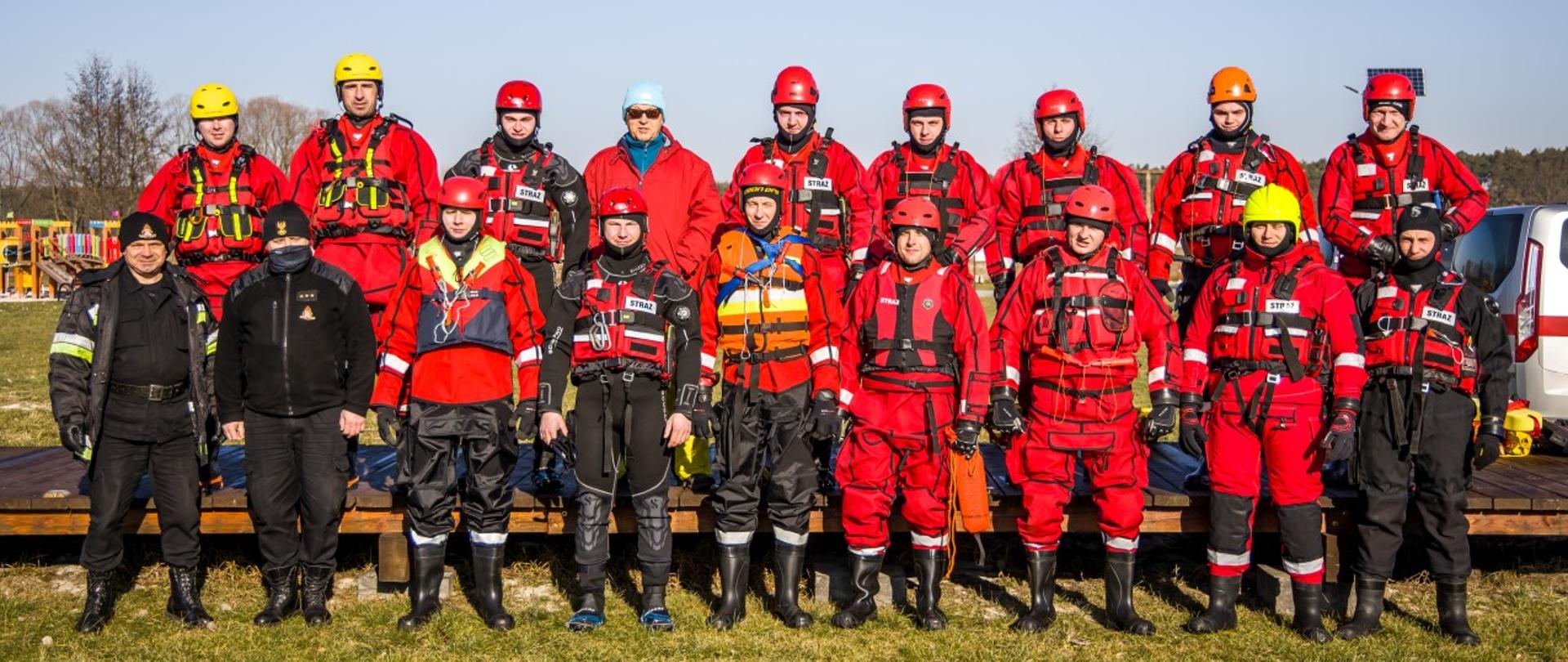 Szkolenie i ćwiczenia z ratownictwa lodowego - wspólne zdjęcie grupowe na miejscu ćwiczeń praktycznych. Strażacy PSP, uczestniczy ćwiczeń w ubraniach wypornościowych i kaskach na głowach.