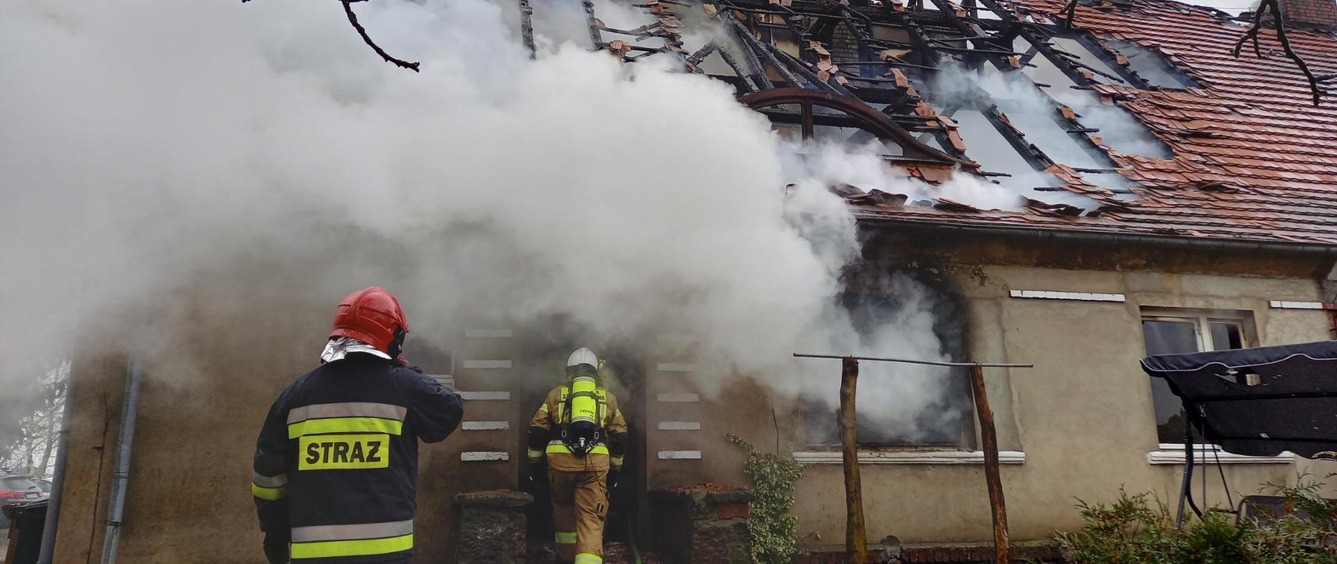 Kłęby dymu wydobywające się z budynku. Widoczny zniszczony dach w tym nadpalone elementy konstrukcyjne. Przed wejściem do budynku dwóch strażaków.