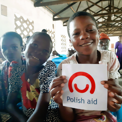 Troje czarnoskórych dzieci pozuje do zdjęcia. Jedno z nich trzyma wydrukowane logo polskiej pomocy.