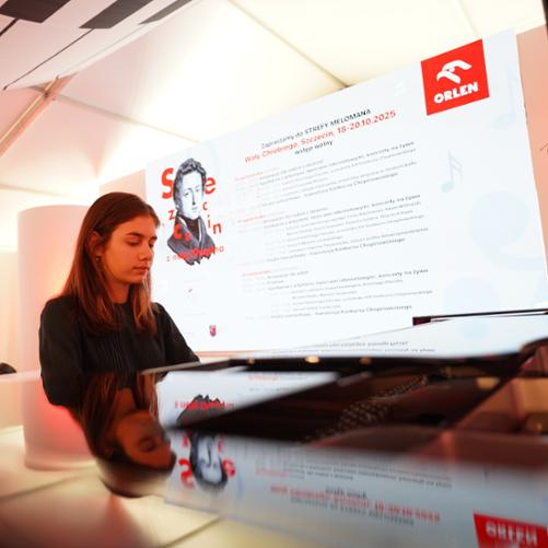 zdjęcie przedstawia uczennicę grającą na fortepianie na tle ekranu multimedialnego z logo sponsora koncertu