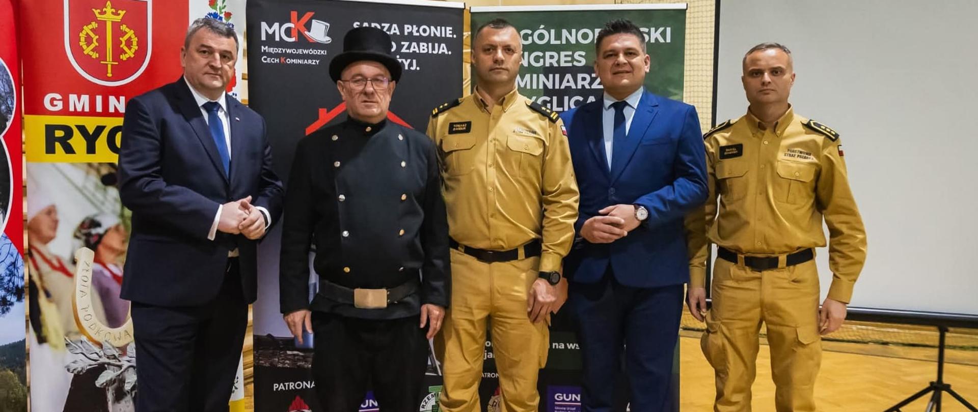 Obok siebie w szeregu stoją mężczyźni, strażacy ubrani są w piaskowe mundury, kominiarz ma czarny uniform na głowie kapelusz, pozostali mężczyźni ubrani są w ciemne garnitury. Z tyłu widać bilbordy. 