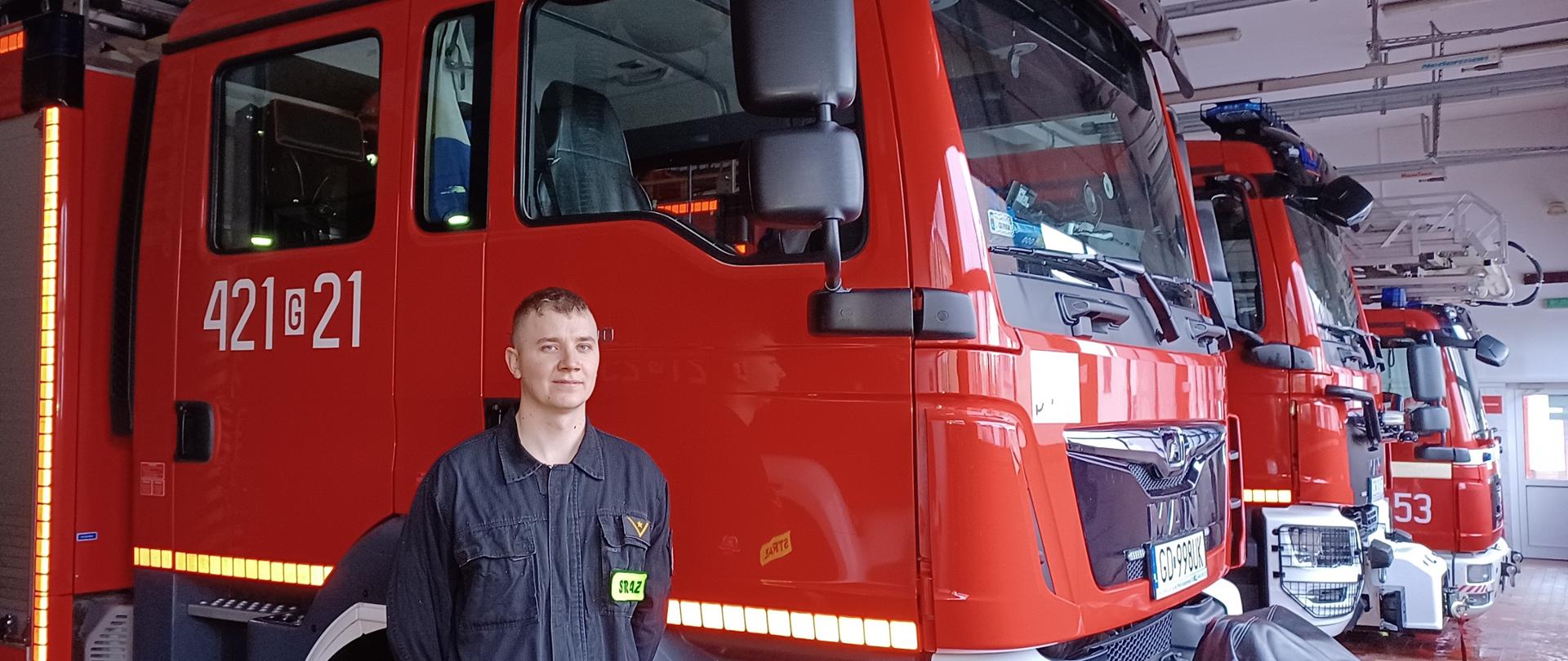 Zdjęcie przedstawia mł. asp. Krystiana Drzewickiego stojącego przy samochodach pożarniczych w pomieszczeniach garażowych Jednostki Ratowniczo – Gaśniczej w Człuchowie