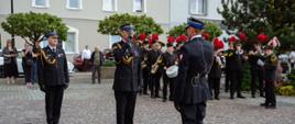 Fotografia przedstawia oficjalną, uroczystą meldunek strażacki na placu miejskim. Trzech oficerów straży pożarnej w mundurach galowych stoi w centrum, a jeden z nich salutuje drugiemu. W tle widać dętą orkiestrę strażacką oraz cywilnych gapiów. Akcja dzieje się w słoneczny dzień na wybrukowanym placu.