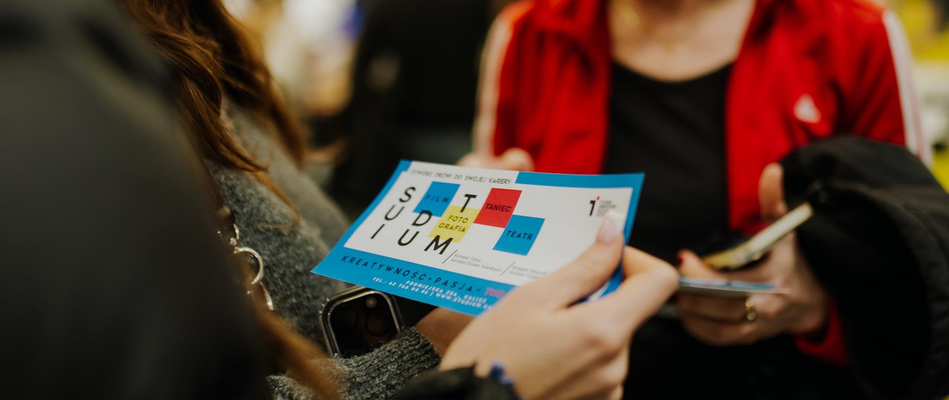 Zbliżenie na ulotkę Studium Animatorów Kultury trzymaną przez uczestniczkę targów. W tle kobieta w czerwonej kurtce prowadzi rozmowę.