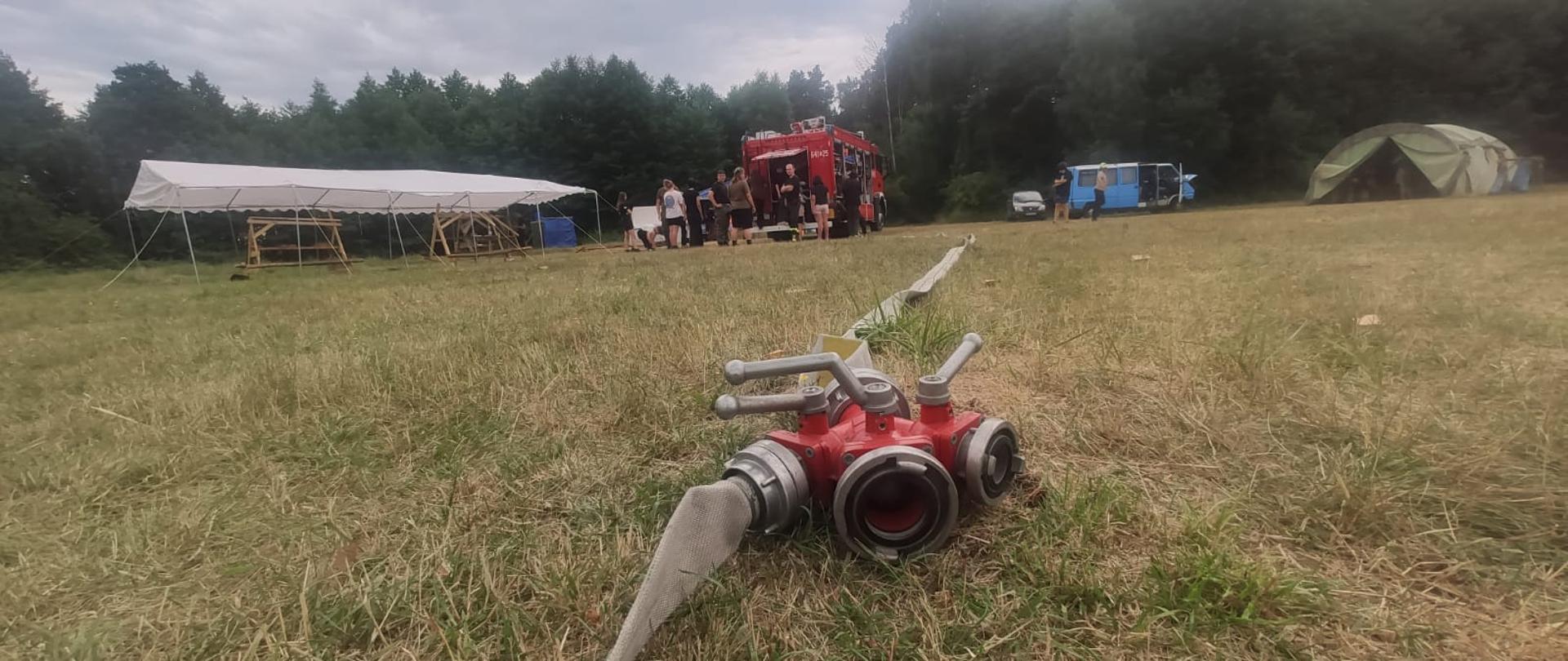 Zdjęcie wykonane w lesie podczas obozu harcerskiego. W tle widoczni harcerze stojący przy wozie strażackim. Na pierwszym planie rozdzielacz z wężem strażackim.