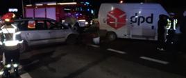 Na zdjęciu widoczne dwa samochody na skrzyżowaniu po kolizji. Przed samochodami znajdują się strażacy, za samochodami wóz strażacki. Zdjęcie w porze nocnej.