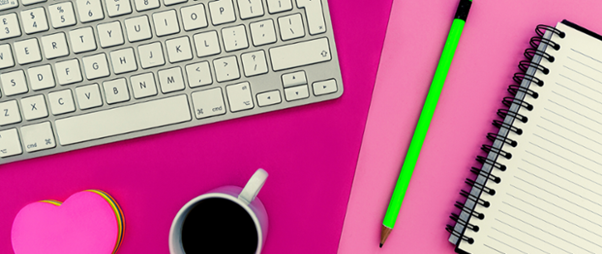 Na zdjęciu widoczna szara klawiatura oraz notes w kształcie serca z kolorowymi kartkami. Obok stoi różowy kubek. Po prawej stronie notes w linie oraz zielony ołówek. Przedmioty leżą na różowym blacie. 