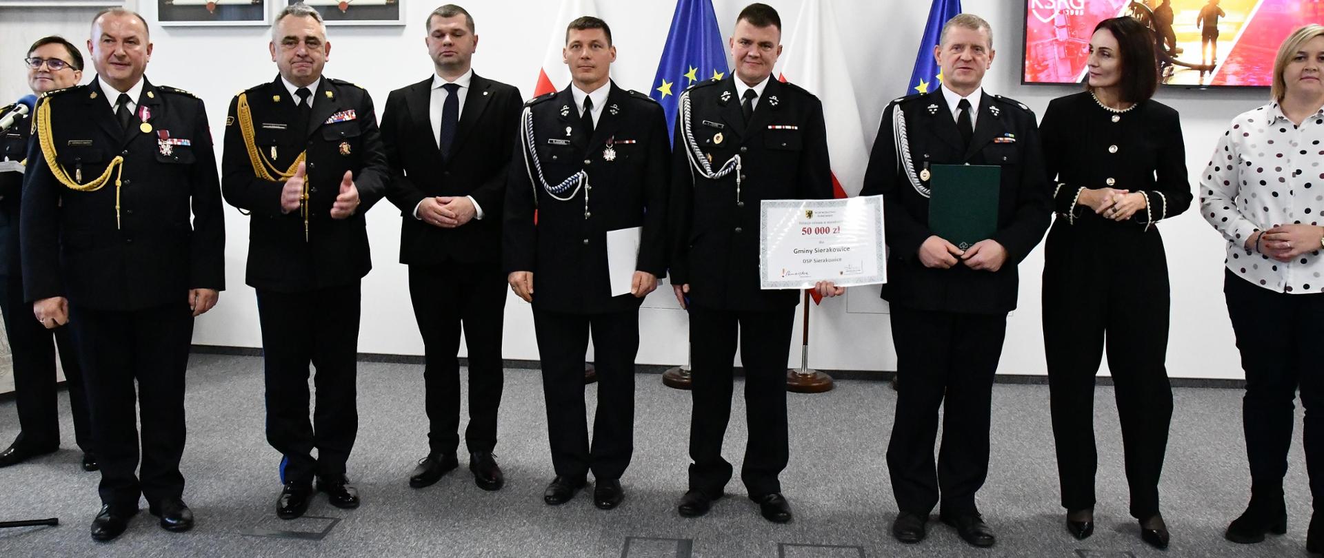 Grupa osób w mundurach galowych i strojach formalnych stoi w rzędzie, jedna osoba trzyma dyplom. W tle flagi Polski i UE oraz ekran z prezentacją PSP.