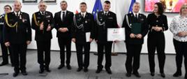 Grupa osób w mundurach galowych i strojach formalnych stoi w rzędzie, jedna osoba trzyma dyplom. W tle flagi Polski i UE oraz ekran z prezentacją PSP.