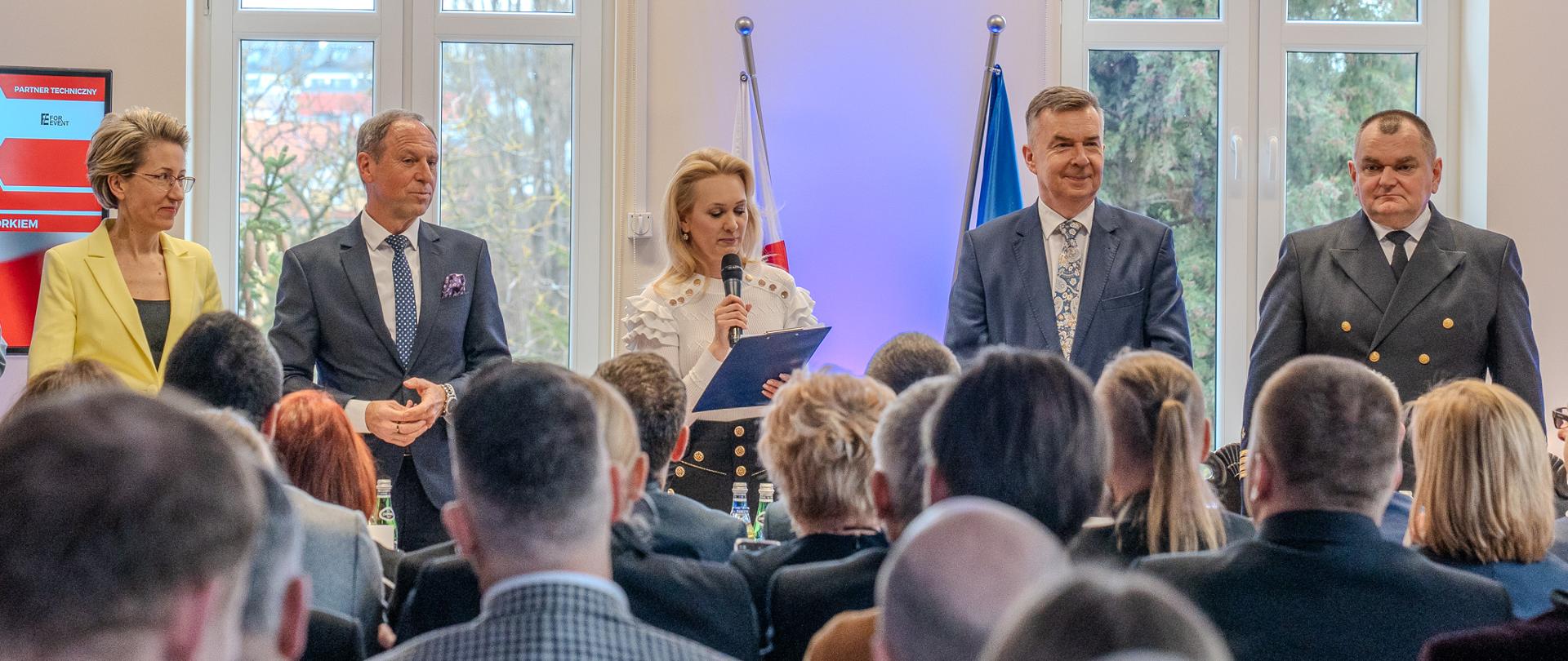 Zdjęcie od tyłu sali, minister Wieczorek i kilka osób stoją nad stołem, naprzeciwko na ustawionych w rzędy krzesłach siedzą ludzie.