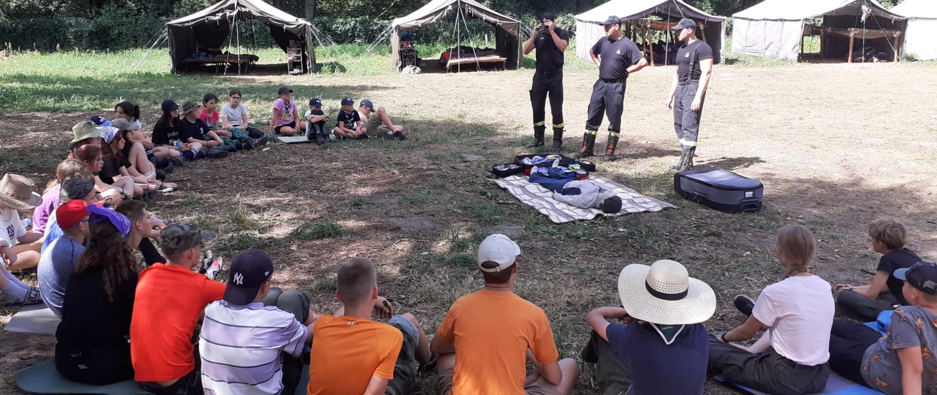 Dzieci siedzą w półokręgu na polanie i słuchają pogadanki trzech strażaków