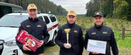 Zdjęcie przedstawia 3 strażaków w ubraniu służbowym. Osoba w środku trzyma puchar za zdobycie drugiego miejsca.
