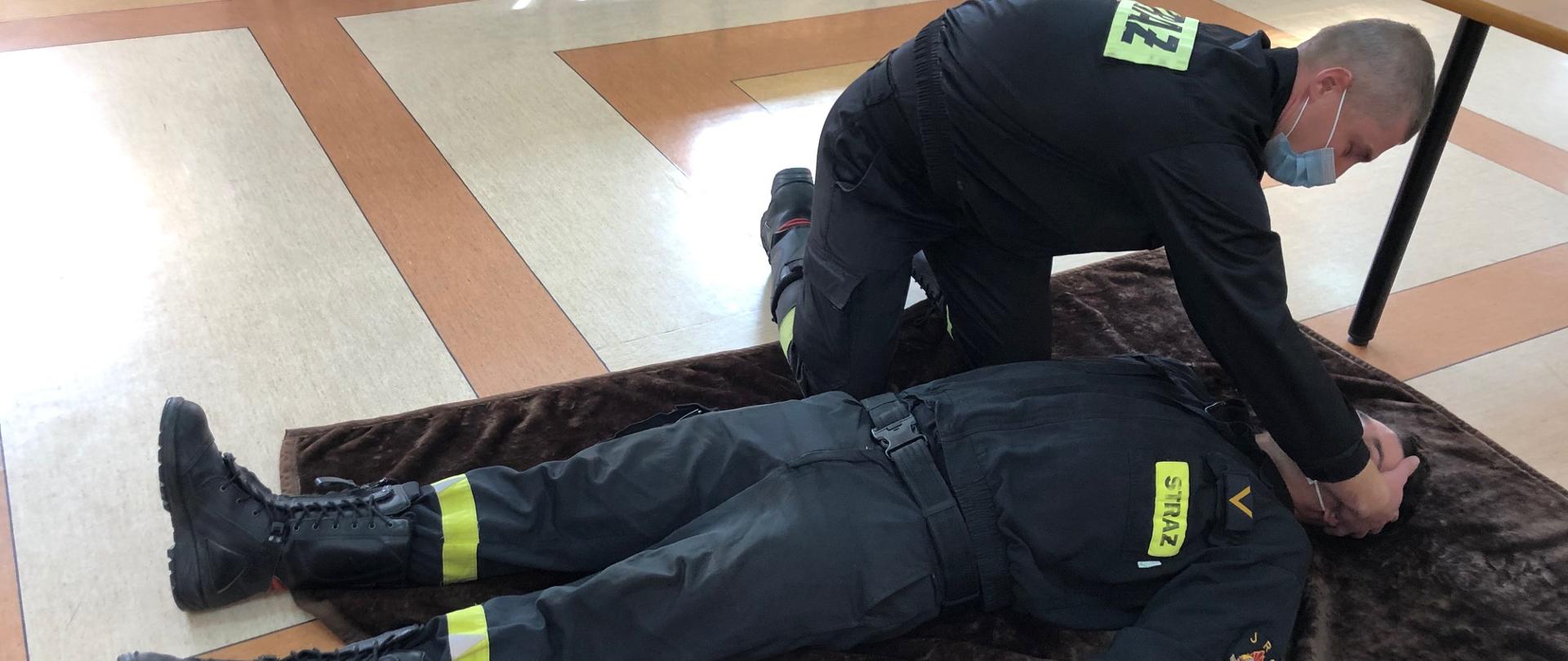 Na zdjęciu strażak- ratownik wykonuje symulację badania urazowego osoby poszkodowanej. Badanie rozpoczyna od głowy, następnie bada szyję, obojczyki, klatkę piersiową, brzuch, miednicę, kończyny dolne, kończyny górne oraz plecy i pośladki. Badanie urazowe polega na szybkim zidentyfikowaniu krwawień oraz innych nieprawidłowości poszczególnych części ciała.