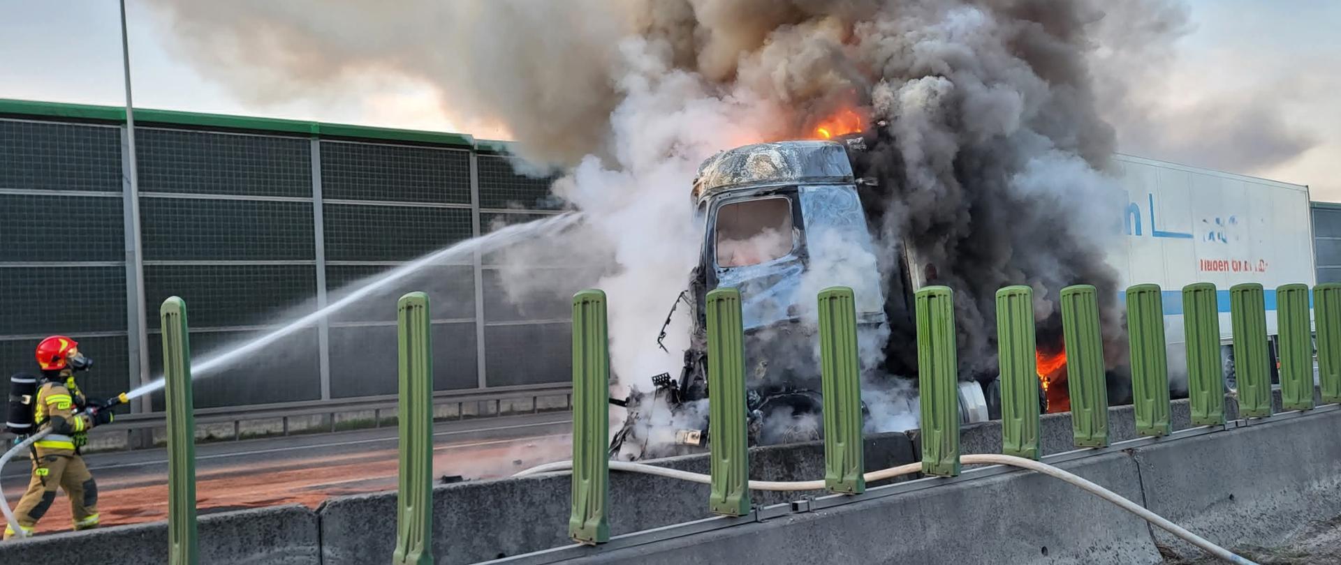 Zdjęcie przedstawia akcję gaśniczą płonącego samochodu ciężarowego na drodze szybkiego ruchu. Z prawej strony kadru widać kabinę ciągnika siodłowego, z której wydobywają się płomienie i gęsty, ciemnoszary dym, który unosi się wysoko w górę. Ogień zajmuje również przednią część naczepy. Po lewej stronie zdjęcia, odwrócony tyłem do obiektywu, stoi strażak w pełnym umundurowaniu ochronnym i hełmie, kierujący strumień wody na płonącą kabinę. Ciężarówka stoi na jezdni, przy barierkach energochłonnych, a za nią widać wysokie ekrany akustyczne. Tło stanowi pochmurne niebo.