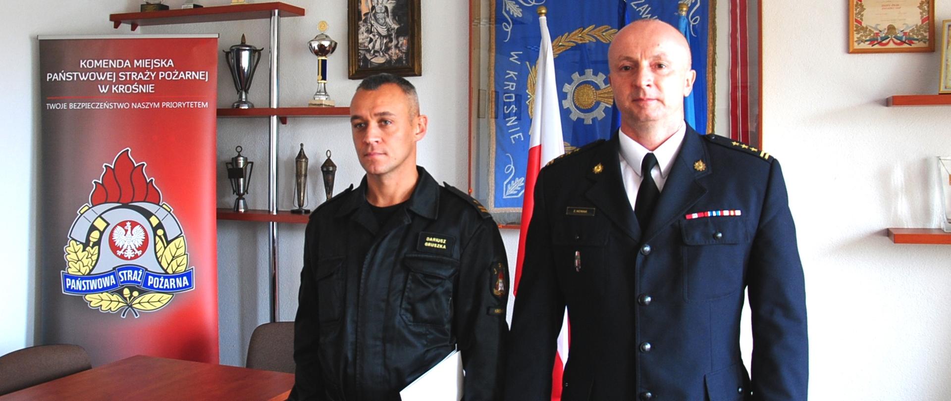 Zdjęcie przedstawia 2 oficerów Państwowej Straży Pożarnej stojących obok siebie w pomieszczeniu świetlicy. W tle, po lewej stronie fotografii widoczny jest czerwony rollup z logotypem Państwowej Straży Pożarnej oraz napisem Komenda Miejska Państwowej Straży Pożarnej w Krośnie. Strażak stojący po prawej stronie ubrany jest w mundur wyjściowy, a strażak po prawej w umundurowanie dowódczo – sztabowe.