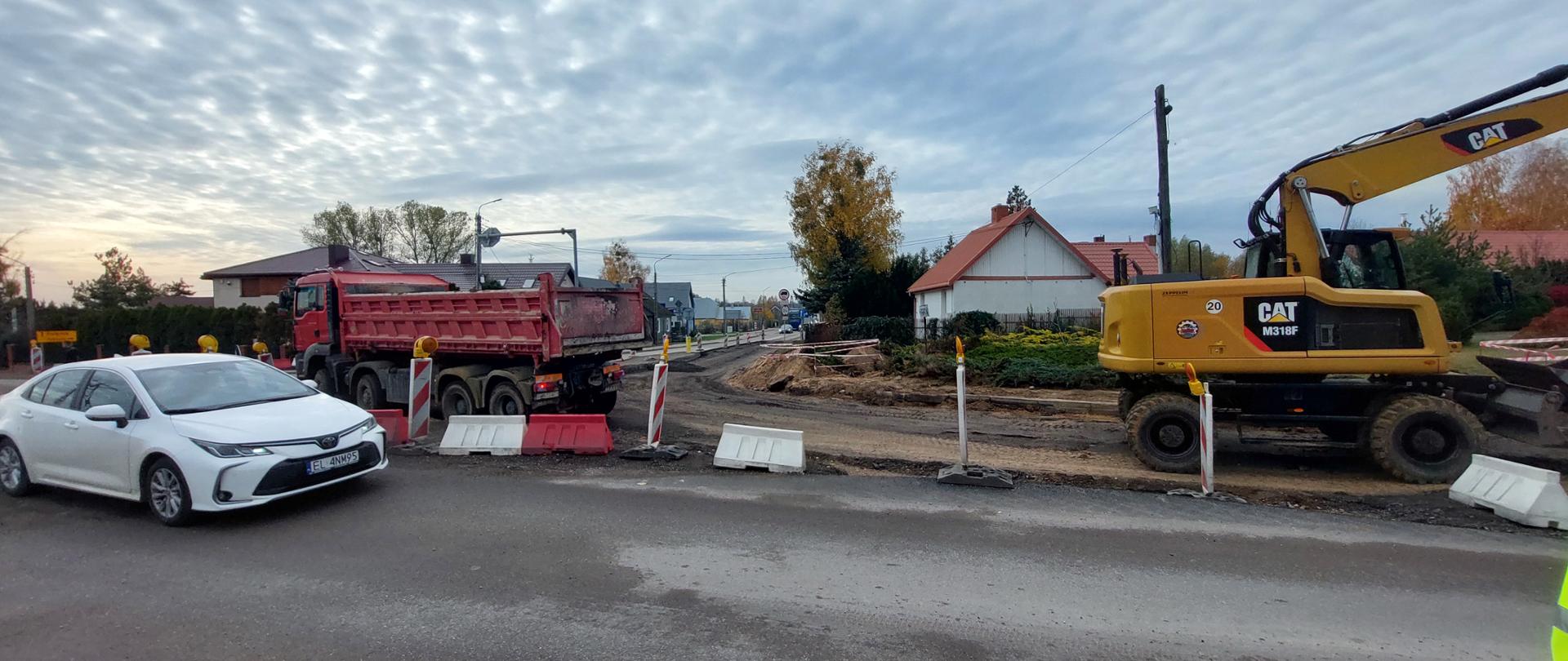Na zdjęciu widać teren budowy drogi. Po lewej stronie stoi biała osobówka, obok niej czerwony samochód ciężarowy z wywrotką. Po prawej znajduje się żółta koparka z wysięgnikiem skierowanym w stronę ziemnych wykopów. Przed maszynami rozstawione są barierki drogowe i oznakowanie tymczasowe. W tle widać kilka domów jednorodzinnych i drzewa, a nad całą sceną rozciąga się niebo pokryte falistymi chmurami.